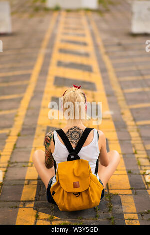 Ragazza moderna si siede con la schiena per la telecamera su una strada di marcatura. Porta un zaino giallo, bracci tatuati Foto Stock