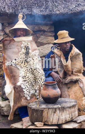 Due Basotho Africano nero uomini seduti nei tradizionali pelli di animali o nasconde al di fuori di una pietra rondawel di paglia o la capanna con una pentola di creta riempito di birra Foto Stock