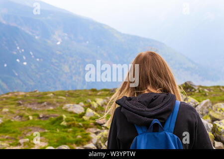 Giovane ragazza bionda viaggiatore con un zaino blu sollevato e godendo il verde paesaggio di montagna. Vista posteriore Foto Stock