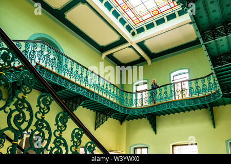 Un visitatore si trova sulla galleria in ferro verde della sala delle scale del Luitpoldbad a Bad Kissingen, in Germania – l'edificio costruito tra il 1868 e il 1871, un tempo il più grande stabilimento termale d'Europa – sotto un lucernario con vetrate colorate che corona la scala. Foto Stock