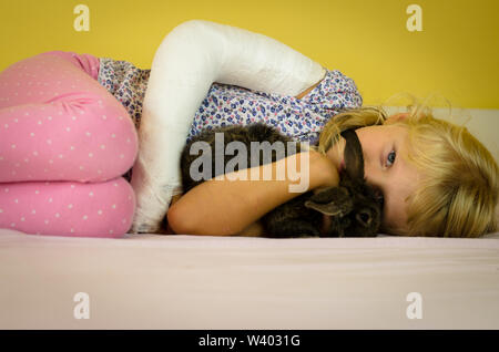 Hurt ragazza bionda con mano rotto giacente sul letto e tenendo bunny rabbit pet Foto Stock