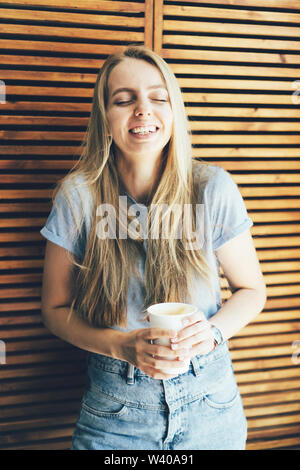 Una studentessa hipster bionda con capelli lunghi ride da un divertente scherzo. Bicchiere di carta con caffè in mano. La gioventù di pausa caffè per momenti felici. Foto Stock