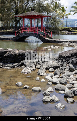 Un rosso ponte coperto al parco in Hilo, Hawaii con costa rocciosa e alberi Foto Stock