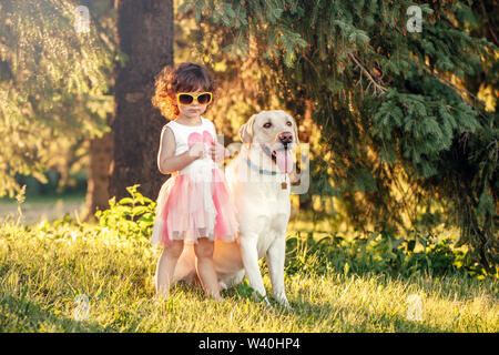 Carino adorabili poco ricci ragazza caucasica indossando occhiali da sole giallo con il suo cane nel parco al di fuori al tramonto sul giorno di estate. Bambini che giocano con sottoprodotti di origine animale non Foto Stock