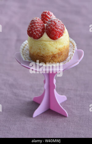 Deliziosi mini Cheesecake ai lamponi e rosa sul cupcake stand su viola pallido sfondo di lino. Il fuoco selettivo sulla parte anteriore di lamponi. Formato verticale. Foto Stock