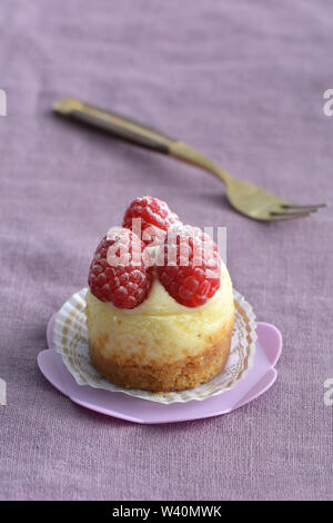 Deliziosi mini Cheesecake ai lamponi e rosa sul cupcake stand su viola pallido sfondo di lino. Il fuoco selettivo sulla parte anteriore di lamponi. Formato verticale. Foto Stock
