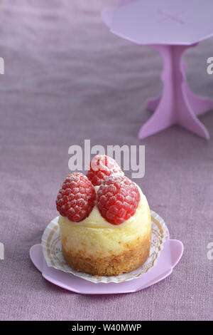 Deliziosi mini Cheesecake ai lamponi e rosa sul cupcake stand su viola pallido sfondo di lino. Il fuoco selettivo sulla parte anteriore di lamponi. Formato verticale. Foto Stock