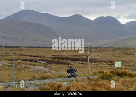 Due persone su una moto sfilacciare su una strada che attraversa la terra della torbiera nella Contea di Galway verso il Connemara con le montagne o Twelve Bens di Connemara avanti. Foto Stock