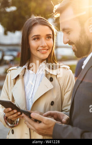 Foto di ottimista lavoratori ufficio uomo e donna in abbigliamento formale sorridente e utilizzando i telefoni cellulari su una strada di città Foto Stock