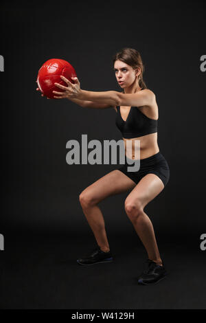 A piena lunghezza Ritratto di skinny donna caucasici in sportswear facendo sit ups con palla fitness durante gli allenamenti in palestra isolate su sfondo nero Foto Stock