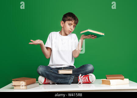 Pensieroso giovane ragazzo con lentiggini scegliendo tra libro e vuoto copyspace mentre guardando la telecamera su sfondo verde Foto Stock