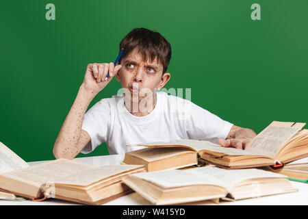 Pensieroso giovane ragazzo con le lentiggini che guarda lontano mentre seduti a tavola con i libri su sfondo verde Foto Stock