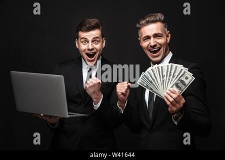 Due felici gli imprenditori eccitati indossando tute in piedi isolato su sfondo nero, tenendo il computer portatile, mostrando il denaro banconote Foto Stock