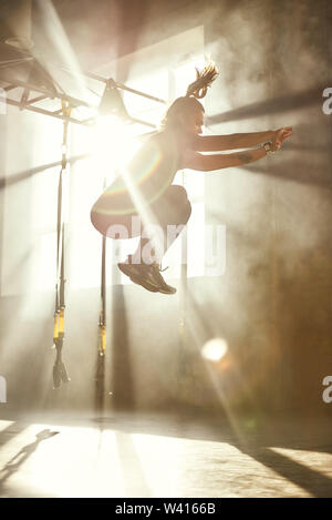 Superiore e superiore. Vista laterale della giovane donna atletica in sport abbigliamento jumping mentre ti alleni in palestra professionale. Sport concept. TRX Training. Allenamento Foto Stock