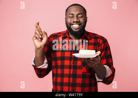 Ritratto di un felice eccitato uomo africano indossando camicia isolata permanente, tenendo la piastra con la torta di compleanno, tenendo le dita incrociate Foto Stock