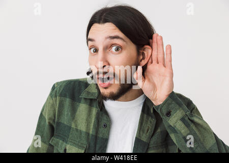 Ritratto di un giovane curioso barbuto brunette uomo che indossa plaid shirt in piedi isolato su sfondo bianco, cercando di ascoltare le voci Foto Stock