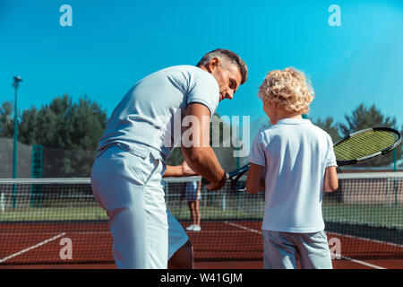 Curly blonde boy ascolto di padre prima di giocare a tennis al di fuori Foto Stock