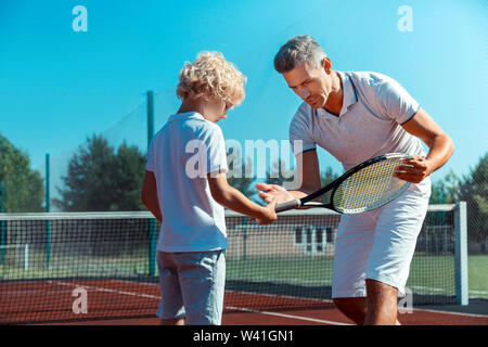 Curly blonde figlio ascoltando padre tenendo racchetta da tennis Foto Stock