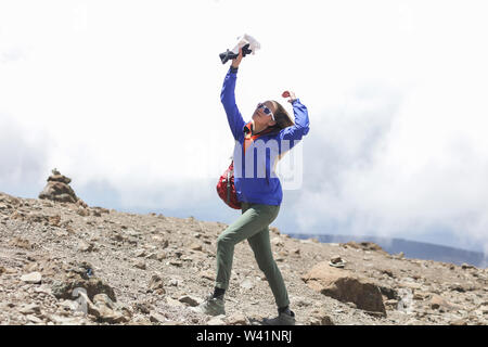 Giovane biondo con stringa corpo shapely salì sulla cima della montagna. Indossando moderno e vestiti comodi, rosso zaino e occhiali da sole. Capelli sventolare Foto Stock