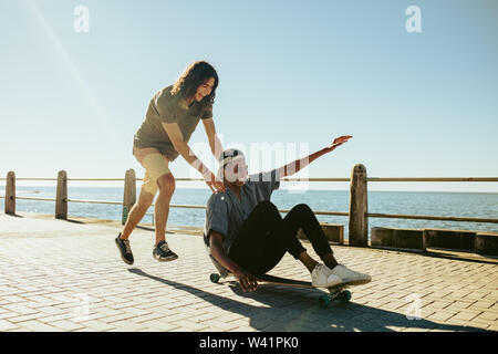 Felice giovane uomo seduto su longboard che viene spinto dal suo amico lungo la strada che dal mare. Ragazzi godendo all'aperto sul lungomare. Foto Stock