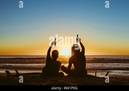Vista posteriore di due giovani surfisti birre di tostatura sulla spiaggia e guardare il tramonto. Giovani uomini partying durante il tramonto sulla spiaggia. Foto Stock