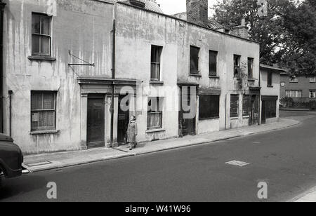 Nei primi anni settanta, storico, una signora camminare lungo uno stretto marciapiede accanto a una vecchia terrazza vittoriana alloggiamento, alcuni abbandonata, Charlton villaggio, a sud di Londra, Inghilterra, Foto Stock