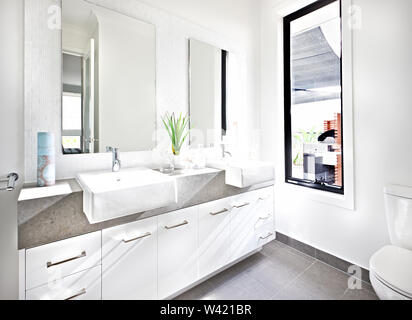 Bagno bianco illuminato con luce solare intorno al contatore dalla finestra in vetro pannello in prossimità dello specchio e impianto di fantasia accanto al lavello e rubinetto sul Foto Stock