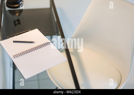 Vista dall'alto di un elegante sedia bianca e un vetro tavolo da studio con un notebook, penna e matita mantenuta su di esso Foto Stock