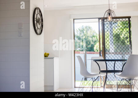Vista di una zona pranzo dotata di splendidi, contemporaneo orologio a muro sulla parete laterale, una luce a soffitto, sedie bianche e un elegante tavolo Foto Stock