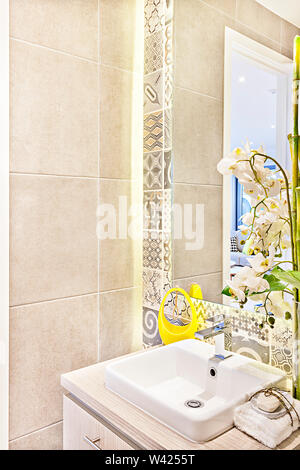 Moderno bagno lavandino e decorativi fiori bianchi accanto a specchio sulla parete con la progettazione del disegno di distribuzione e la luce gialla, la parete di piastrelle da sinistra sid Foto Stock