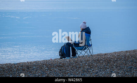 Una giovane donna seduta su una sedia su una spiaggia peeble come lavorare in remoto con il suo computer portatile Foto Stock