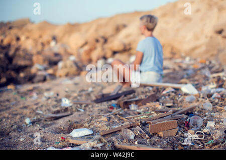 Donna sedersi sulla spiaggia tra i rifiuti. Inquinamento ambientale concetto Foto Stock