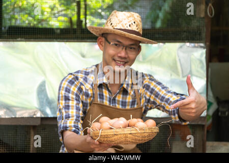 Giovane agricoltore smart usura plaid camicia a maniche lunghe grembiule marrone sono holding fresco uova di pollo nel cestello in una fattoria di pollo in lui home area Foto Stock