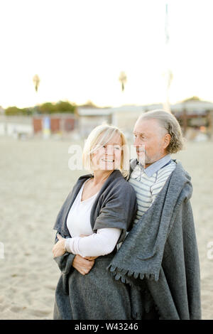 Senior marito caucasica abbracciando la bionda moglie indossando plaid e permanente sulla spiaggia di sabbia. Concetto di felice coppia di anziani. Foto Stock