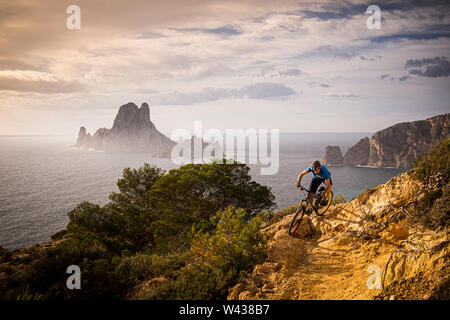 Un uomo corse in mountain bike lungo un sentiero roccioso sulla costa occidentale di Ibiza. Es Vedrá isola e del Mare Mediterraneo sono in background. Foto Stock