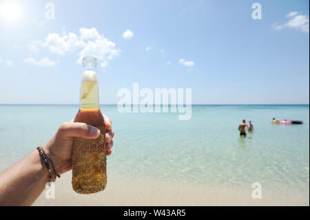 Maschio di mano che tiene una bottiglia di birra contro un cielo sereno ed un mare limpido e cristallino. In background alcune persone avendo divertimento nell'acqua. Concetto di vacanza. Foto Stock