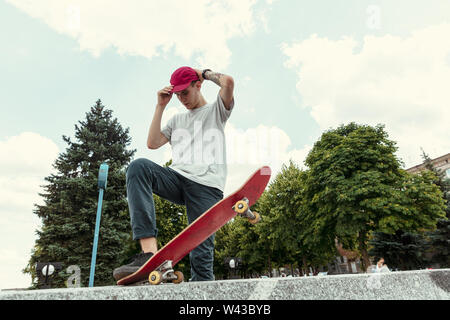 Guidatore di skateboard facendo un trucco presso la città della strada nella giornata di sole. Giovane uomo in sneakers e cappuccio di equitazione e surf su asfalto. Concetto di attività per il tempo libero, sport, extreme, hobby e movimento. Foto Stock