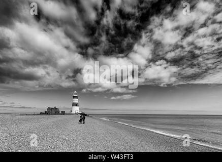 Due persone che camminano su una spiaggia di ciottoli con un faro in background. Foto Stock