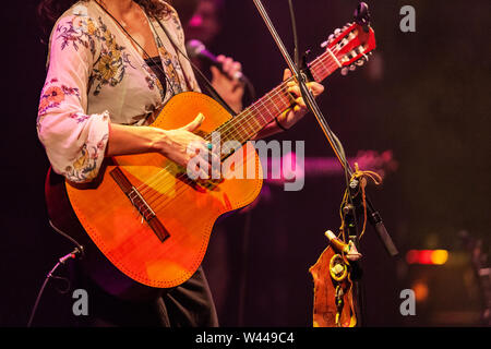 Una vista ravvicinata su un elegante signora membro della band di suonare una chitarra durante un live set di musica. Musicisti sfocate sono visti in background. Dettagli sul palco a gig. Foto Stock