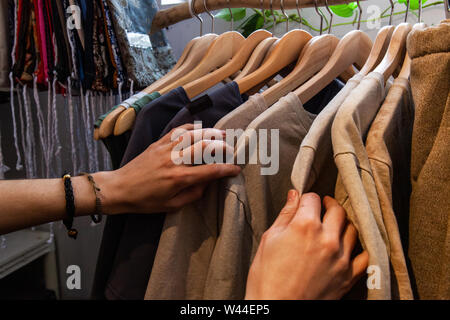 Le mani di una ragazza alla moda sono visti closeup, navigazione abbigliamento racks all'interno di un eco-negozio di moda. Autentica appendere vestiti da un rustico di rampa. Foto Stock