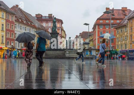 Graz/Austria - Giugno 22, 2019: Persone con ombrello passando da Erzherzog Johann fontana al Hauptplatz (piazza principale) in un giorno di pioggia Foto Stock