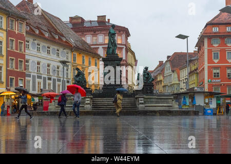 Graz/Austria - Giugno 22, 2019: Persone con ombrello passando da Erzherzog Johann fontana al Hauptplatz (piazza principale) in un giorno di pioggia Foto Stock