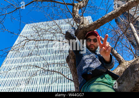 Un basso angolo di visione di un giovane maschio caucasico seduti in un centro città tree, vestito di rosso con fascia verde e pantaloni, che mostra a dita a simboleggiare il p Foto Stock