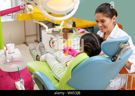 Sorridendo gentile dai capelli scuri dentista donna tenendo un mirror Foto Stock