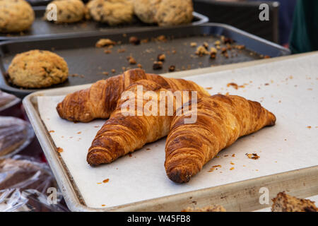 Un primo piano di tre croissant appena sfornati su un vassoio in un mercato in stallo, scones sfocate sono visti sullo sfondo, cibi caldi serviti a una fiera all'aperto. Foto Stock