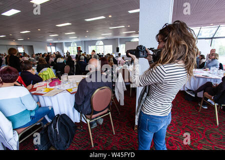 Una femmina di fotografo è visto scattare fotografie durante un montaggio professionale in una grande sala, co-lavoratori raccogliere per eventi aziendali. Foto Stock