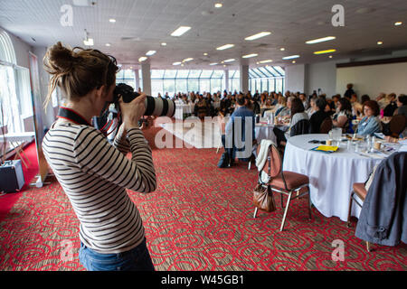 Un oltre la spalla di una donna di scattare la foto nel corso di una assemblea generale, sfocata la gente seduta ai tavoli sono visti in background con copy-spazio Foto Stock