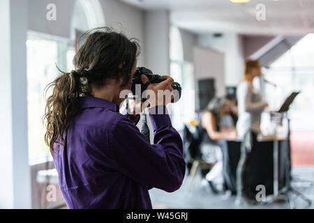 Un oltre la spalla di un fotografo professionista scattare istantanee durante un evento di networking per colletti bianchi, sfocata altoparlante è visto in ba Foto Stock