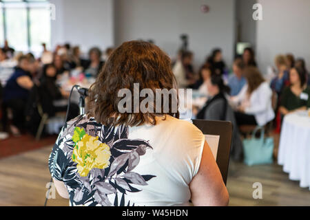 Un sovrappeso donna che indossa un top floreale è visto da dietro, stando in piedi sul palco durante un team building evento. Fiducioso nella parte anteriore di una stanza affollata di colleghi Foto Stock