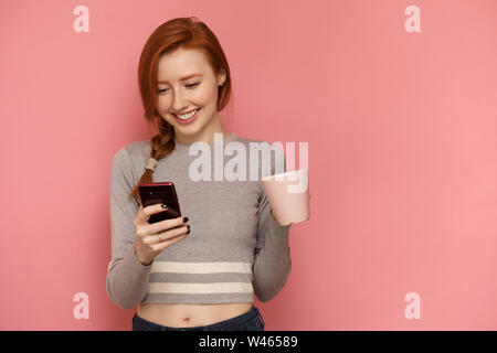 I Capelli rossi ragazza detiene una tazza in una mano guardando il suo smartphone in altra parte e sorridente Foto Stock